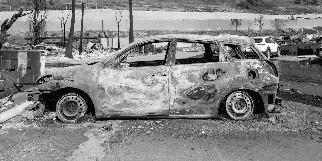 A burned-out hatchback shell with melted paint and no glass, parked on a street